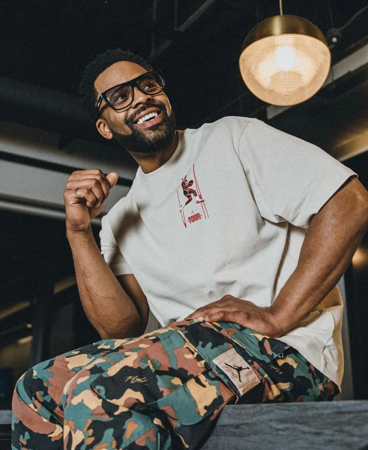 LaRoyce Hawkins laughing, seated in a white T-shirt and camouflage pants